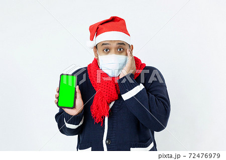 Portrait young asian man wearing medical face mask to protect covid-19, corona virus and blue sweater, red santa hat, red scarf showing smartphone with blank green touch screen on the white background 72476979