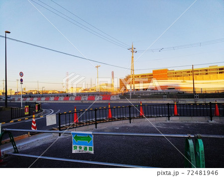 工事中の歩道の向こうにフェンスと車道。送電線の向こうに夕陽に照らされた工事中の建設現場 工事中の歩道の向こうにフェンスと車道。送電線の向こうに夕陽に照らされた工事中の建設現場 72481946