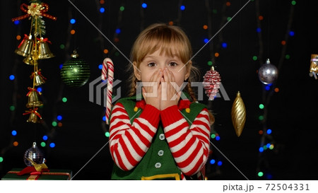 Kid girl in Christmas elf Santa Claus helper costume looks at camera makes big eyes covering mouth Kid girl in Christmas elf Santa Claus helper costume looks at camera makes big eyes covering mouth 72504331