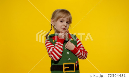 Shy shamed child girl in Christmas elf Santa helper costume posing looking camera, blows air kiss Shy shamed child girl in Christmas elf Santa helper costume posing looking camera, blows air kiss 72504481