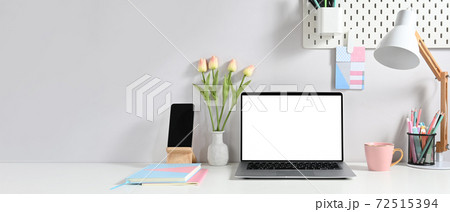 A white blank screen laptop is putting on a white working desk surrounded by various equipment. A white blank screen laptop is putting on a white working desk surrounded by various equipment. 72515394