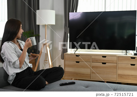 A young woman is sitting on sofa watching tv while holding a cup of coffee in hands. A young woman is sitting on sofa watching tv while holding a cup of coffee in hands. 72515721