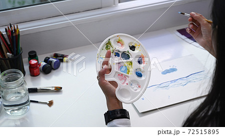 Close up view of female artist holding colour palette and paint brush working in her workshop. Close up view of female artist holding colour palette and paint brush working in her workshop. 72515895