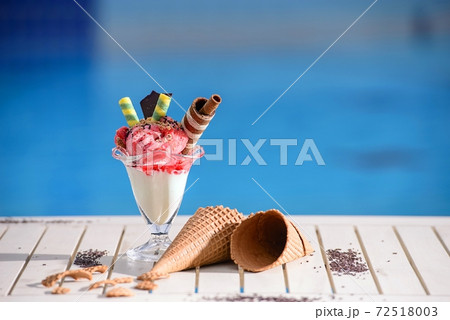 Front view of glass with strawberry ice cream on blur bokeh background with copy space. Raspberry sorbet with waffle chocolate cones on white table top of blue water pool 72518003