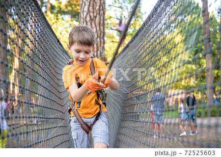 Young cute child boy in summer clothing, safety harness and helmet attached with carbine to cable moves slowly along rope way on green trees sunny bokeh background. Sport, game, leisure concept 72525603