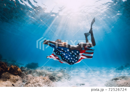 Woman free diver glides underwater over sandy sea with US flag. 72526724