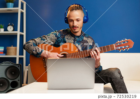 Young man sitting in his room and watching guitar tutorial on laptop 72530219