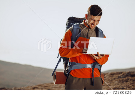 Young man backpacker using his laptop in mountain journey Young man backpacker using his laptop in mountain journey 72530829