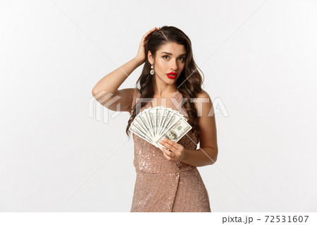 Shopping concept. Beautiful and successful woman in elegant dress, fixing hairstyle and holding money, looking perfect, standing over white background 72531607