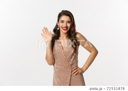 Christmas party and celebration concept. Happy beautiful woman in glamour dress saying hello, smiling and waiving hand with friendly expression, white background 72531876