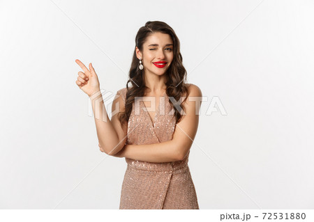Christmas, holidays and celebration concept. Beautiful caucasian woman in party dress and red lipstick, winking and smiling, pointing finger left at promo offer, white background 72531880