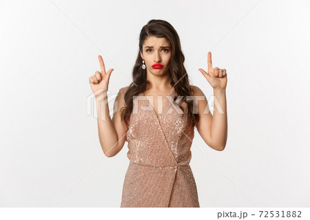 New Year, christmas and celebration concept. Gloomy and sad glamour girl in party dress pointing fingers up, sulking and crying about something, standing over white background New Year, christmas and celebration concept. Gloomy and sad glamour girl in party dress pointing fingers up, sulking and crying about something, standing over white background 72531882