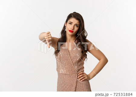 Skeptical young woman in elegant dress looking disappointed, showing thumbs-down, dislike christmas party, standing over white background Skeptical young woman in elegant dress looking disappointed, showing thumbs-down, dislike christmas party, standing over white background 72531884