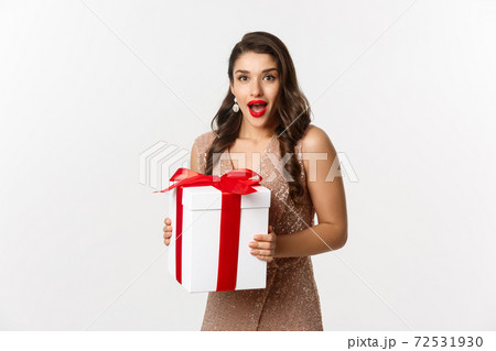 Merry Christmas. Image of beautiful woman in glamour dress receiving gift and looking surprised, celebrating New Year, standing over white background 72531930