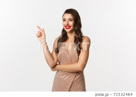 Christmas, holidays and celebration concept. Attractive woman wearing party dress and red lipstick, pointing and looking left, enjoing new year eve, white background Christmas, holidays and celebration concept. Attractive woman wearing party dress and red lipstick, pointing and looking left, enjoing new year eve, white background 72532464