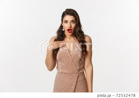 Winter holidays and party concept. Image of attractive woman with red lips, wearing glamour dress and pointing at herself with shock, looking surprised, standing over white background 72532478