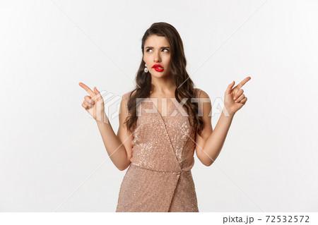 Christmas party. Thoughtful woman in glamour dress pointing fingers sideways, making choice, thinking and showing left and right offers, standing over white background Christmas party. Thoughtful woman in glamour dress pointing fingers sideways, making choice, thinking and showing left and right offers, standing over white background 72532572