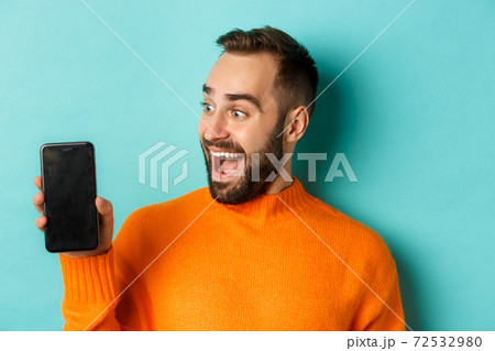 Close-up of young bearded man showing phone screen and looking amazed, wearing orange sweater, standing against studio background Close-up of young bearded man showing phone screen and looking amazed, wearing orange sweater, standing against studio background 72532980