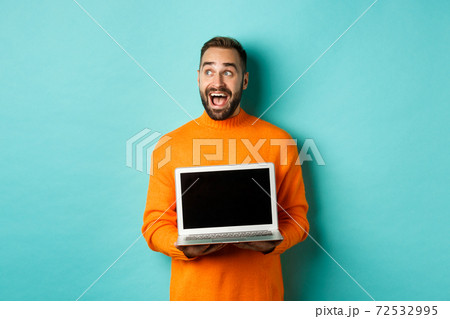 Amazed young man in orange sweater looking at upper left corner, showing laptop screen promo offer, standing over turquoise background Amazed young man in orange sweater looking at upper left corner, showing laptop screen promo offer, standing over turquoise background 72532995