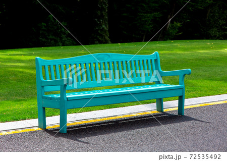 A blue wooden bench lit by sunlight stands on an asphalt road with yellow markings in a park with a green lawn and large trees in the background. A blue wooden bench lit by sunlight stands on an asphalt road with yellow markings in a park with a green lawn and large trees in the background. 72535492