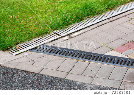 the intersection of the asphalt road and the pedestrian sidewalk made of tiles with the grating of the drainage system in the park with green grass. the intersection of the asphalt road and the pedestrian sidewalk made of tiles with the grating of the drainage system in the park with green grass. 72535501