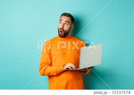 Handsome man doing online shopping, looking up thoughtful while using laptop, standing over light blue background Handsome man doing online shopping, looking up thoughtful while using laptop, standing over light blue background 72537578