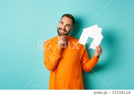 Real estate concept. Handsome adult man in orange sweater thinking about buying flat or renting, holding house maket and looking thoughtful, standing over blue background Real estate concept. Handsome adult man in orange sweater thinking about buying flat or renting, holding house maket and looking thoughtful, standing over blue background 72538116