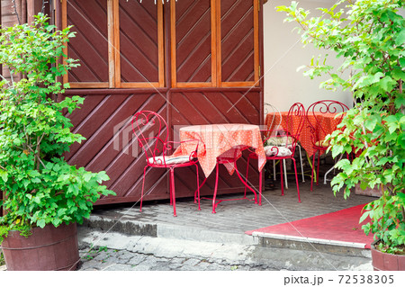 a free red iron table with chairs and a polka-dot tablecloth in an outdoor cafe by the front of the building with wooden elements and green leafy plants, nobody. a free red iron table with chairs and a polka-dot tablecloth in an outdoor cafe by the front of the building with wooden elements and green leafy plants, nobody. 72538305