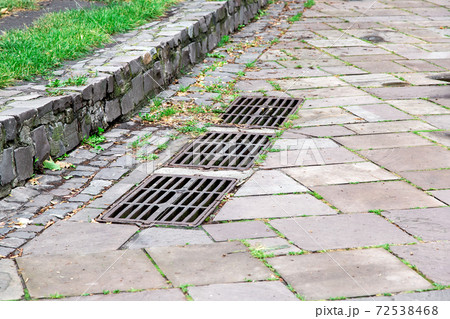the iron grate of the drainage system hatch on the footpath paved with paving stones and green grass, close up. the iron grate of the drainage system hatch on the footpath paved with paving stones and green grass, close up. 72538468