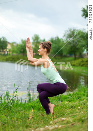 Slender Caucasian brunette girl doing yoga in summer on a green lawn by the river. Eagle Pose, Garudasana Slender Caucasian brunette girl doing yoga in summer on a green lawn by the river. Eagle Pose, Garudasana 72538621