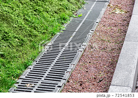 drainage system on the side of the road behind a border of pebbles and gutter with an iron grate and green grass. 72541853