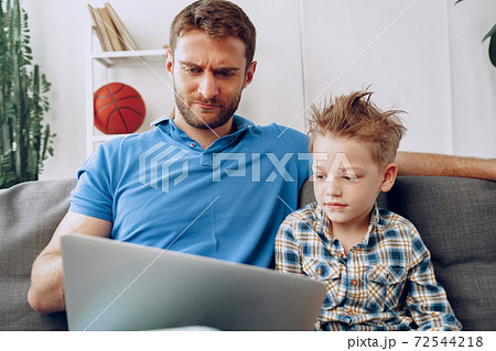 Father and son sitting on couch and watching something on laptop 72544218