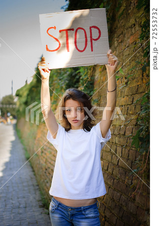 Young protesting woman in white shirt and jeans holds protest sign broadsheet placard with slogan 'Stop' for public demonstration on wall background. 72553732
