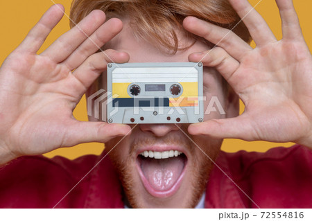 Close up of young ginger man in red jacket holding a cassette Close up of young ginger man in red jacket holding a cassette 72554816