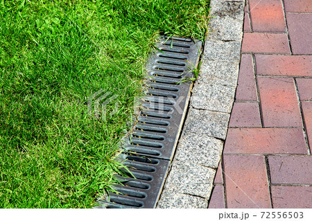 iron gutter with grate to the drainage system on the side of the pedestrian walkway with green lawn on summer day in well maintained park. iron gutter with grate to the drainage system on the side of the pedestrian walkway with green lawn on summer day in well maintained park. 72556503