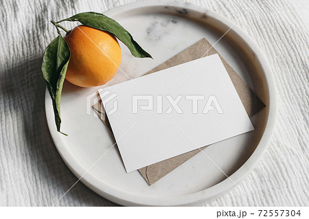 Summer wedding stationery mock-up scene. Blank greeting card, invitation. Craft envelope and orange fruit on elegant marble tray. White muslin tablecloth background. Top view. 72557304
