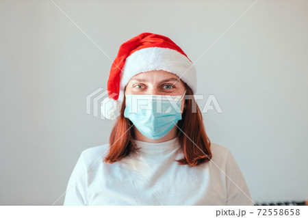 Close up of girl in santa hat and protective medical white mask on grey wall background. New Year in quarantine during the coronavirus pandemic 72558658