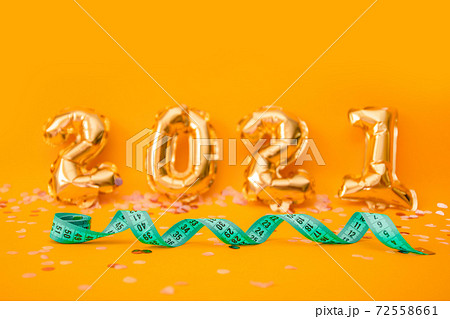 Green measuring tape or tape measure on orange background from 2021 foil balloons with confetti. Preparation for celebrating new year or christmas, weight loss, fitness concept 72558661