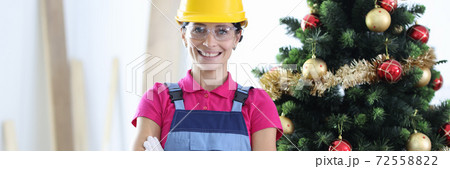 Woman in yellow protective helmet and construction overalls smiling in office near new year tree Woman in yellow protective helmet and construction overalls smiling in office near new year tree 72558822