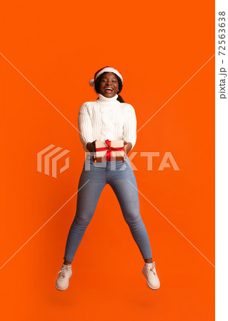 Cheerful black woman in santa hat jumping with christmas present in hands 72563638