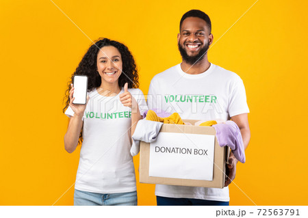 Two Volunteers Holding Donation Box Showing Phone Screen, Yellow Background Two Volunteers Holding Donation Box Showing Phone Screen, Yellow Background 72563791