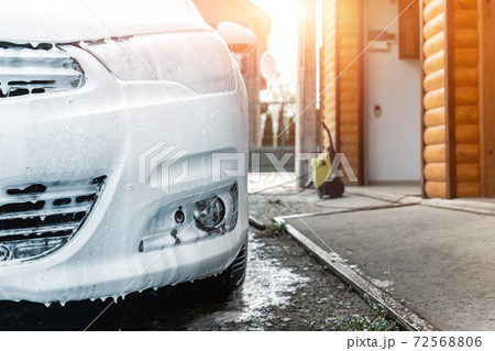Close-up detail car wash with high pressure water equipment pump at home backyard outdoor on bright shiny summer day. Vehicle covered with foam shampoo chemical detergents during carwash self service 72568806