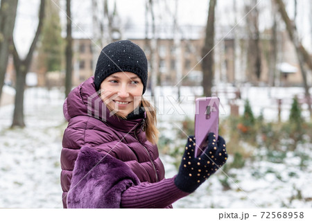 Young happy woman in warm purple dawn jacket smiling while making selfie with smartphone during walk in winter city park. Pretty girl enjoys winter vacations and travel 72568958
