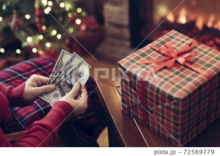 Woman putting american dollars in red christmas sock sitting near christmas tree and fireplace and packing gift box. Woman putting american dollars in red christmas sock sitting near christmas tree and fireplace and packing gift box. 72569779