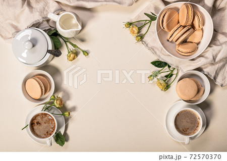 Frothy coffee in white cups, delicious French macaroons. Dessert still life on a beige background. 72570370