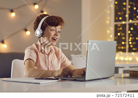 Schoolboy in headphones answers a teacher's question while sitting at home in front of a laptop. Schoolboy in headphones answers a teacher's question while sitting at home in front of a laptop. 72572411