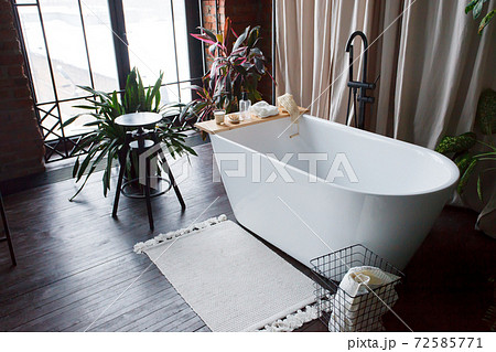 Modern bathroom with freestanding white acrylic bathtub and black loft mixer. Walls made of bricks, big window, parquet is wooden on floor 72585771