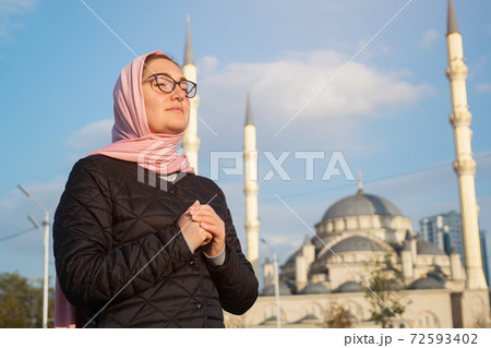 Young woman in a pink scarf and glasses whispers something while holding her hands on the background of the mosque Young woman in a pink scarf and glasses whispers something while holding her hands on the background of the mosque 72593402