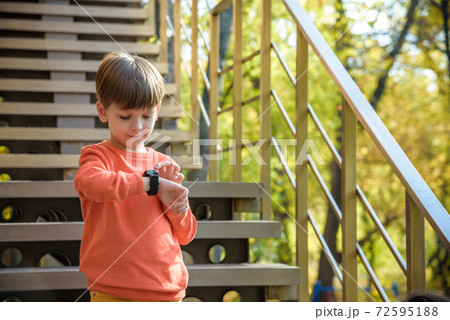 A cute boy wearing stylish shirt stay near school, looking at his smart watch touching the screen. A child using electronic device. Lifestyle concept 72595188
