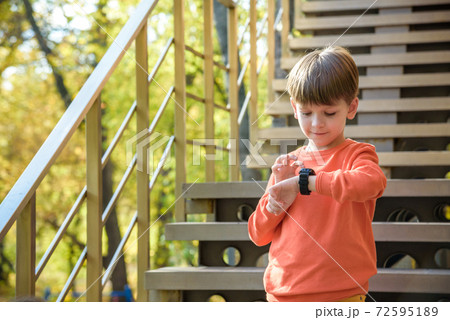 A cute boy wearing stylish shirt stay near school, looking at his smart watch touching the screen. A child using electronic device. Lifestyle concept 72595189